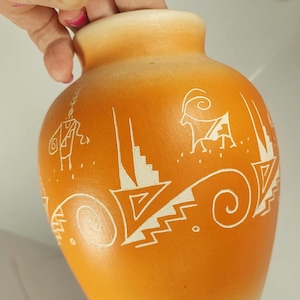 May include: Orange ceramic vase with white tribal designs. The vase has a rounded body and a narrow neck. The white designs include geometric shapes, spirals, and stylized animal figures. The vase is held by a hand with pink nail polish.