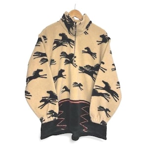 May include: Beige fleece pullover featuring a black horse design. The top has a high collar, a quarter-zip, and a black and red patterned hem. The long-sleeved top is suspended from a wooden hanger.