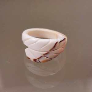 Peut inclure: Une bague sculptée avec un motif de feuille. La bague est principalement blanche avec des accents marron et beige. Le design de la bague présente des lignes sculptées qui créent un motif en forme de feuille. La bague est faite d'un matériau lisse et poli.