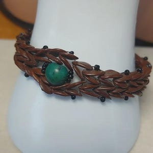 May include: A brown bracelet with a woven design, featuring a central turquoise bead and small black beads. The bracelet is displayed on a white surface, showcasing its intricate details and craftsmanship.