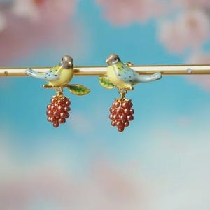 May include: A pair of earrings with bird figures in blue and yellow, perched on a gold-coloured branch. Each bird has a cluster of red beads hanging below. The earrings are designed with a nature-inspired theme, suitable for everyday wear.