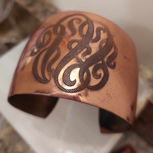 May include: A copper cuff bracelet with an engraved monogram. The monogram is in a script font and is centred on the bracelet.