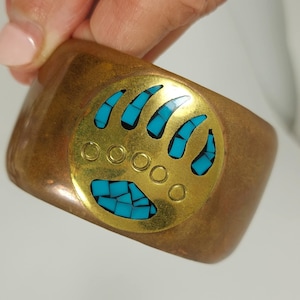 Op de afbeelding: Een brede, koperkleurige manchetarmband. De armband heeft een goudkleurig cirkelvormig middelpunt met een berenpootontwerp. De berenpoot is ingelegd met turkooiskleurige stenen. De armband wordt door een persoon vastgehouden.