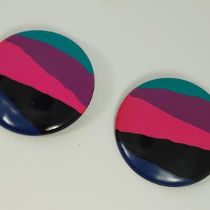 May include: A pair of round, colorful earrings with a layered design. The earrings are made of black, blue, pink, purple, and teal.