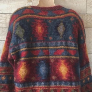 Puede incluir: Un suéter holgado y acogedor en una vibrante mezcla de colores. El diseño presenta un patrón repetido de diamantes, triángulos y formas florales en tonos de rojo, azul, amarillo y verde. El suéter tiene cuello redondo y mangas largas.