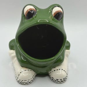 May include: A glossy green ceramic frog figurine with a wide, open mouth. The frog has large, expressive eyes with white and brown details. It is wearing white shoes with black stitched detailing on the front, creating a hollow container shape.