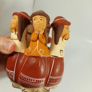 May include: A ceramic figurine depicting multiple figures with brown hair and hats, dressed in orange and white attire. The base is reddish-brown with white decorative patterns. The figurine is held by a hand.
