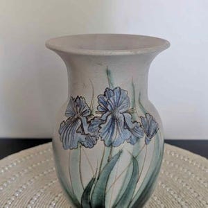 May include: A ceramic vase in light grey, decorated with blue iris flower illustrations and green leaves. The vase has a wide mouth and a flared shape. The vase is on a round, beaded placemat.