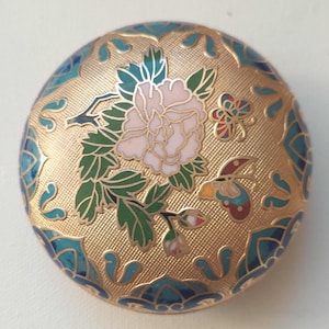 May include: A circular, gold-coloured decorative item with a floral and butterfly design. The central flower is pink with green leaves, and the butterflies have red, blue, and yellow accents. The border features blue and gold patterns.