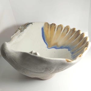 May include: A ceramic bowl with a shell-like shape and a matte white exterior. The interior features a smooth white surface accented by a vibrant blue streak and a series of radiating tan and brown ridges along the scalloped rim edge.