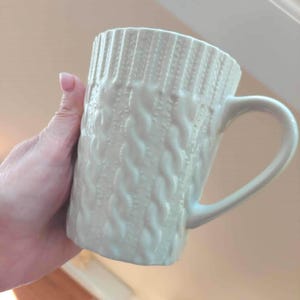 May include: A cream-coloured ceramic mug featuring a raised, textured cable knit pattern across its entire exterior surface. The mug has a smooth, curved handle on the right side and a cylindrical shape with a slightly flared rim at the top.