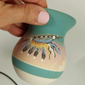 May include: A small, teal-colored ceramic vase with a pink and white painted design of a Native American headdress with feathers.
