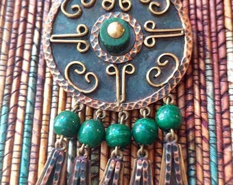 May include: A circular brooch featuring a dark, hammered copper frame with gold-toned wire scrollwork and a central green stone. Five green beads hang from the bottom, each attached to a dangling, hammered copper, bell-shaped metal charm.