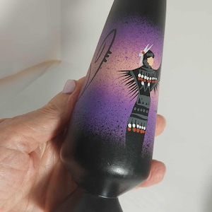 May include: Black vase with a Native American design. The vase has a black base and a purple and black upper section with a painted figure in traditional attire. The figure is adorned with white feathers and a patterned garment.