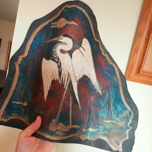 May include: A piece of irregular-shaped leather featuring a hand-painted illustration of a white heron with black markings. The background is a textured blend of deep blue, teal, and reddish-brown tones, framed by a thin, metallic gold border.