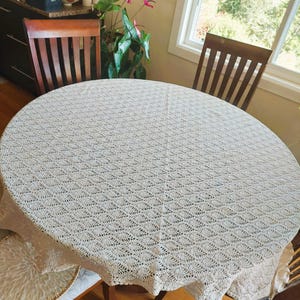 May include: A round, off-white crochet tablecloth featuring a repeating leaf-like pattern throughout the fabric. The textile is draped over a circular table, showcasing the intricate, open-work design and the delicate texture of the yarn.