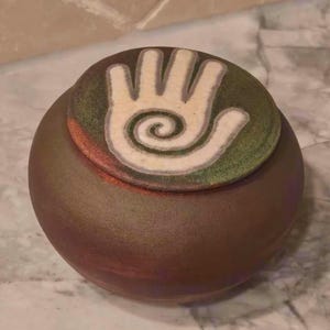 May include: A round, dark brown ceramic jar with a matching lid. The lid features a circular, textured green surface with a prominent, light-colored handprint design in the center, which incorporates a spiral pattern within the palm area.