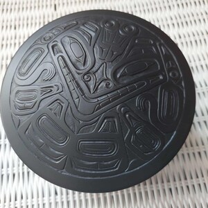 Puede incluir: Un objeto negro circular que presenta un diseño en relieve profundamente tallado en su superficie superior. El patrón consiste en formas geométricas estilizadas e interconectadas y líneas fluidas que forman una composición compleja y simétrica en la tapa.