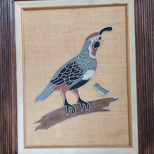 May include: A framed wall art piece featuring a stylized quail and a smaller bird perched on a branch. The artwork is rendered in shades of gray, black, and reddish-brown on a textured tan background, enclosed within a dark brown wooden frame.
