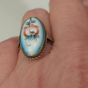 Könnte beinhalten: Ovale Ring mit floralem Design. Der Ring hat einen weißen Hintergrund mit einer handgemalten orangefarbenen und blauen Blume. Der Rand des Rings ist aus verdrehtem Silbermetall. Der Ring befindet sich an einem Finger.