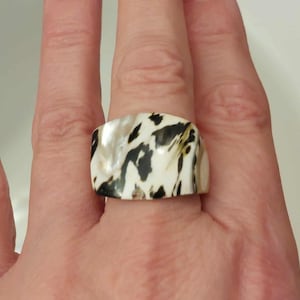 Peut inclure: Une bague large à la surface polie présentant un motif organique marbré dans des tons crème, blanc et brun foncé. Le matériau possède un éclat irisé lisse, affichant des stries sombres irrégulières et fluides sur la base claire.