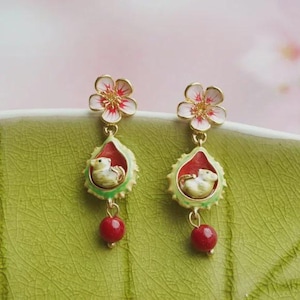 May include: A pair of gold-toned earrings featuring a floral design with red and white petals. Each earring has a teardrop-shaped pendant with a small animal figure inside, accented with red and green enamel, and a red bead at the bottom.