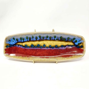 Puede incluir: Un plato de servir de cerámica con un diseño abstracto colorido. El plato tiene una base marrón con un diseño amarillo, rojo, azul y verde.