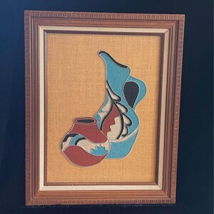 May include: A framed artwork featuring two stylized pottery vessels, one large and one small, with a blue, red, black, and white design on a beige burlap background.