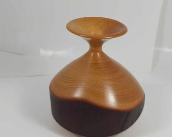 May include: A handcrafted wooden bowl with a unique design. The bowl features a light brown upper section and a dark brown lower section. The bowl has a wide, shallow top and a rounded base. The wood grain is visible, adding to its natural aesthetic.