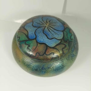 Könnte beinhalten: Eine runde Holzschatulle mit einer blauen Blume, die auf den Deckel gemalt ist. Die Schachtel hat eine irisierende Oberfläche in Grün, Blau und Braun.