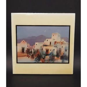 May include: A square ceramic tile featuring a Southwestern landscape painting. The artwork depicts adobe buildings with a mountain backdrop under a dusky sky. The tile has a cream-colored border and a black frame around the painting.