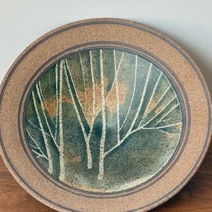 Puede incluir: Plato de cerámica redondo con diseño de árbol. El plato presenta un borde marrón y un diseño central de siluetas de árboles blancos sobre un fondo verde y marrón. El plato es probablemente hecho a mano.