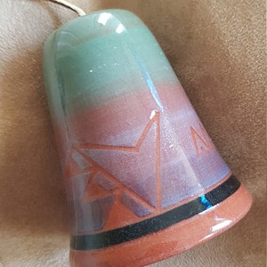 May include: A small, ceramic bell with a green, pink, and brown glaze. The bell has a black band around the rim and a geometric design on the front.