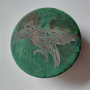 May include: Round, green decorative piece with a silver-colored bird design. The bird is stylized with intricate details, set against a mottled green and brown background. The piece has a rustic, handcrafted appearance.