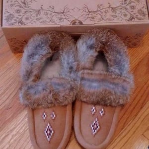 May include: A pair of tan suede moccasin slippers featuring a thick brown and grey fur trim around the opening. The top of each slipper is decorated with a small beaded diamond pattern. The slippers are positioned in front of a cardboard box.