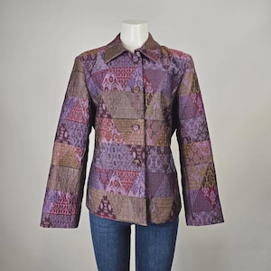May include: A long-sleeve button-up jacket featuring a patchwork design of triangular fabric panels in shades of purple, lavender, and bronze. The garment has a structured collar, a front button closure, and is paired with blue denim jeans.