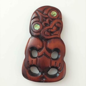May include: A dark brown wooden pendant carved into a stylized anthropomorphic figure with two circular cutouts in the torso and two smaller cutouts near the base. The figure features two inlaid iridescent green abalone shell circular eyes.