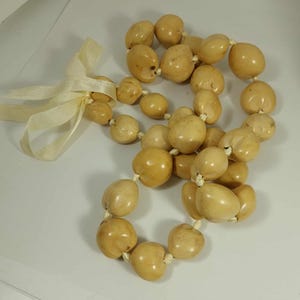 May include: A necklace composed of large, light-colored, rounded beads. The beads are strung together with a white string. A cream-colored ribbon is tied at one end of the necklace, forming a bow. The beads have a smooth, glossy finish.