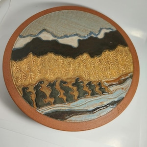 May include: A circular ceramic plate with a landscape design. The plate features a textured forest, mountains, and a river in shades of brown, tan, blue, and grey. The rim is a reddish-brown colour.