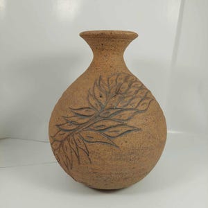 Puede incluir: Un jarrón de cerámica marrón hecho a mano con una superficie texturizada. El jarrón presenta un diseño de hojas en relieve en la parte delantera. El jarrón tiene un cuerpo redondeado y un cuello acampanado, adecuado para exhibir flores o como pieza decorativa.