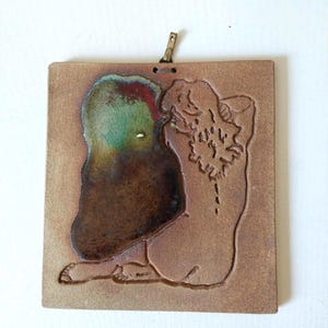 May include: A square ceramic wall plaque featuring a textured tan surface with a debossed outline of a seated figure. The figure is partially filled with a glossy, multi-coloured glaze in shades of deep brown, green, and red. A metal hook is attached.
