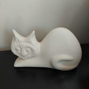 May include: A matte white ceramic cat figurine in a resting position. The sculpture features a smooth, unglazed surface with moulded details defining the ears, facial features, and a curved tail tucked against the body in a continuous form.