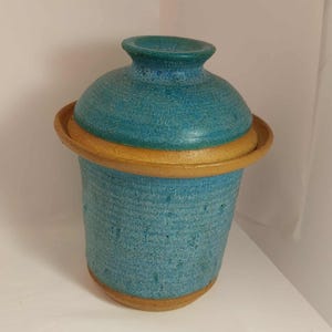 May include: A turquoise ceramic jar with a lid. The jar has a textured surface and a contrasting tan band around the middle and base. The lid matches the jar's turquoise hue. The handmade jar is a decorative item, suitable for storage or display.