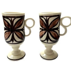 Peut inclure: Deux tasses à café en céramique blanche avec des motifs géométriques bruns et noirs. Les tasses ont un piédestal et une poignée.