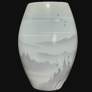 May include: A smooth, oval-shaped ceramic vase featuring a soft, matte finish in shades of pale grey and white. The surface displays a monochromatic landscape illustration of layered mountain ranges, evergreen trees, and birds in flight.