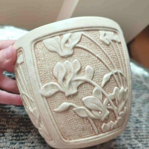May include: A cream-coloured ceramic planter featuring a raised relief design of stylised flowers and leaves. The surface has a textured, grid-like background pattern behind the floral motifs, with a subtle crackle glaze finish throughout the piece.