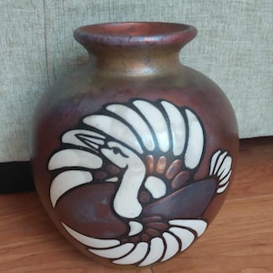 May include: A round ceramic vase with a metallic copper and bronze finish. The front features a large, stylized white bird graphic with dark brown accents. The vase has a wide, curved neck and a smooth, reflective surface throughout the body.
