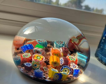 May include: A clear glass paperweight with a dome shape containing a collection of colorful millefiori glass canes. The interior features various small floral patterns in shades of blue, green, orange, yellow, and red embedded in a copper-toned base.
