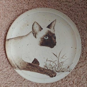 May include: A round, off-white ceramic plate featuring a Siamese cat illustration. The cat is depicted in shades of brown and cream, with blue eyes. The plate has a speckled texture and a simple grass design.