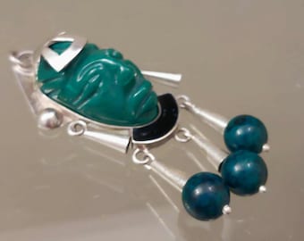 May include: A silver pendant featuring a carved green stone face mask, a black semi-circular accent, and three dangling silver cones tipped with round, dark teal mottled beads. The metal components have a polished finish, and the design incorporates a loop at the top for attachment to a chain or cord.